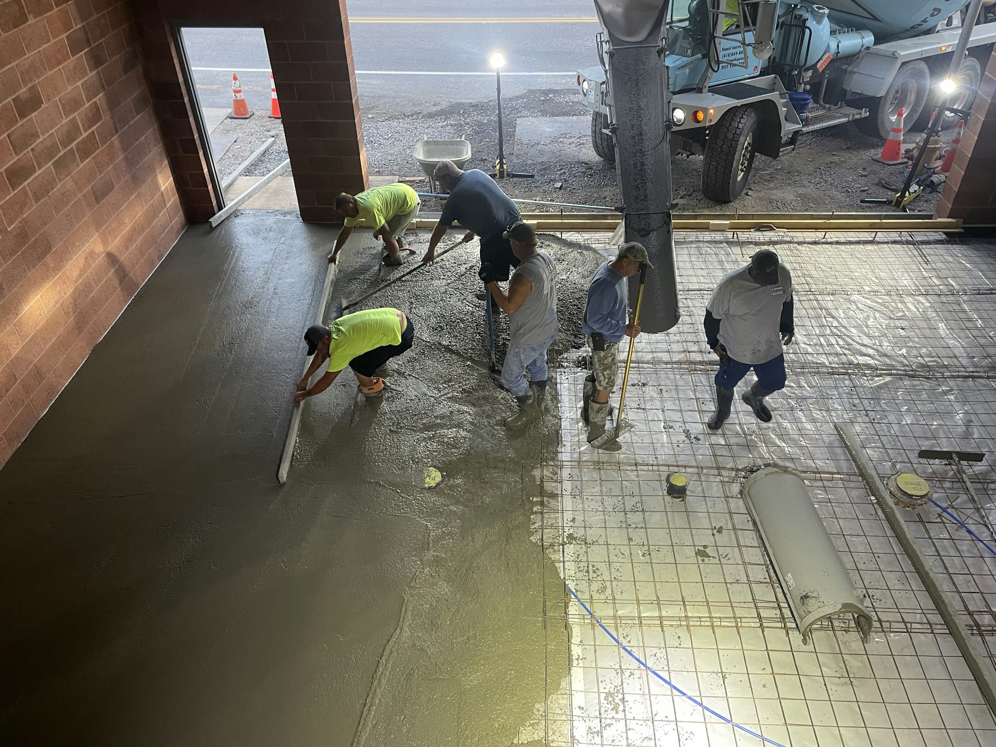 Workers are smoothing wet concrete near a concrete truck, with construction cones visible outside.
