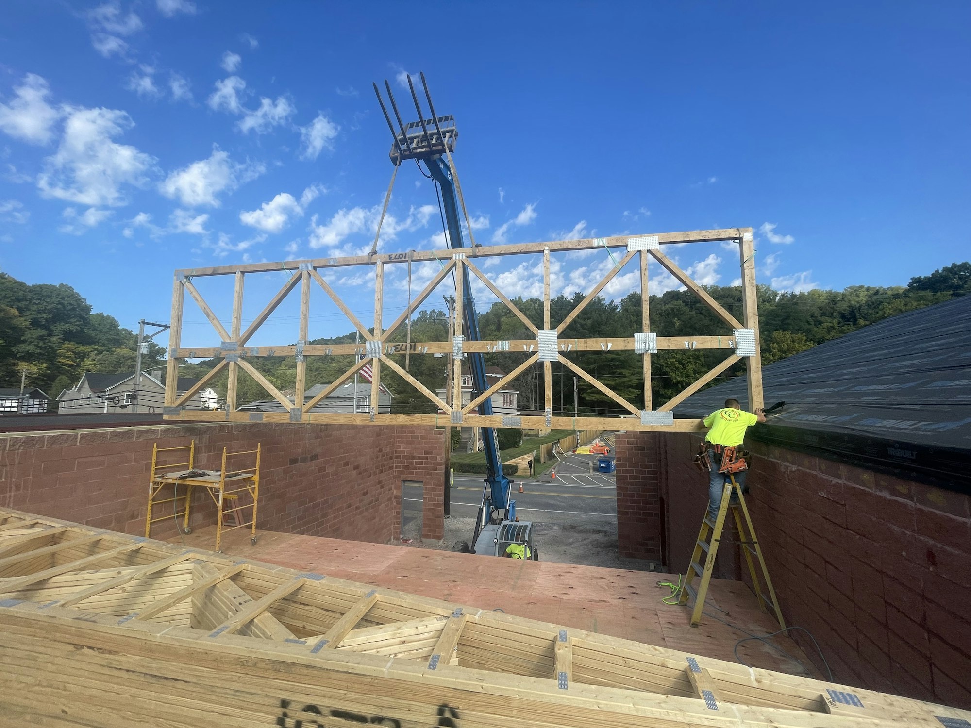 Construction site with wooden trusses being installed, a worker on a ladder, bricks, and a crane against a blue sky.