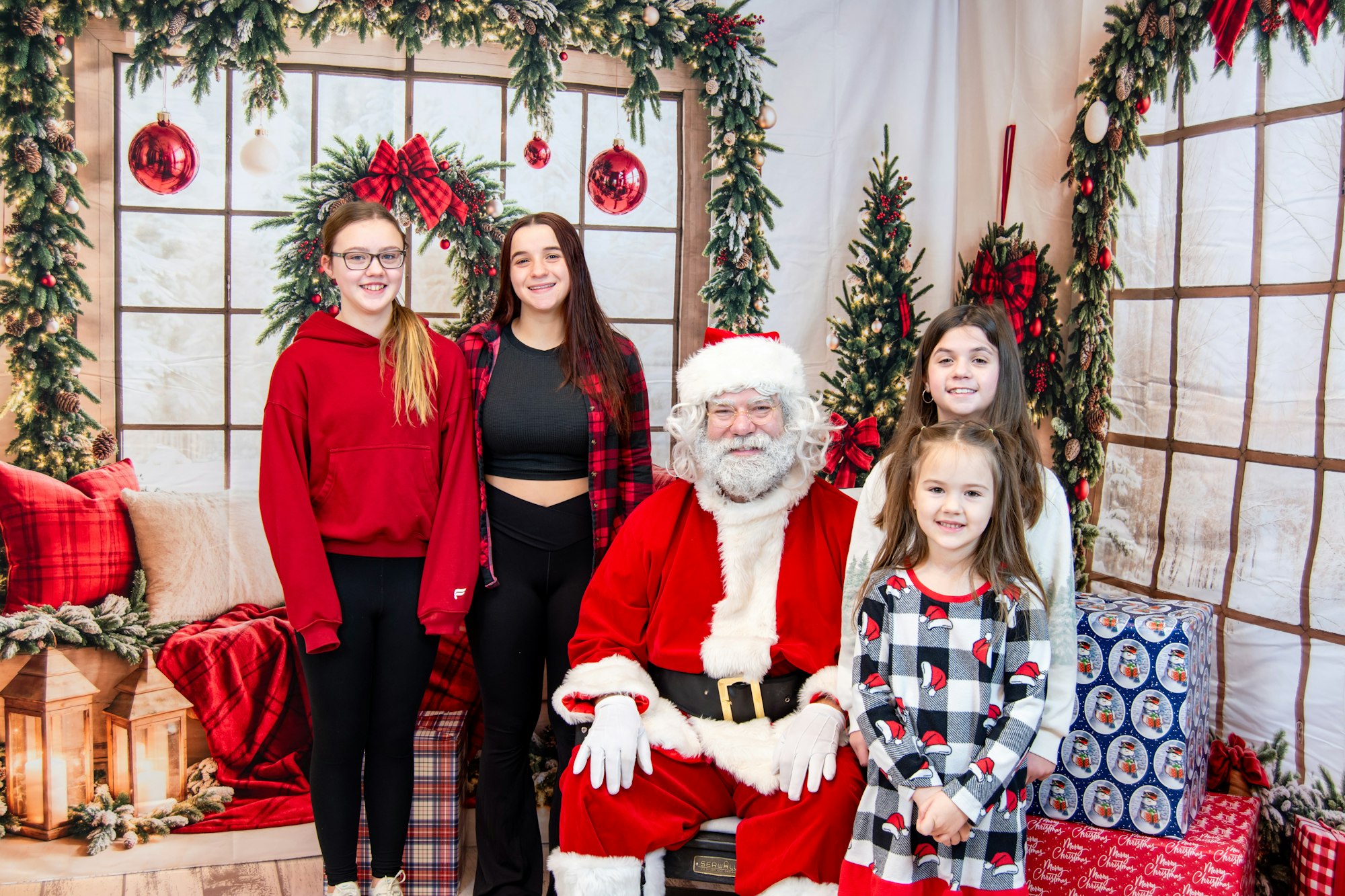 A festive scene featuring Santa Claus surrounded by four girls, set against a holiday backdrop with decorations and gifts.