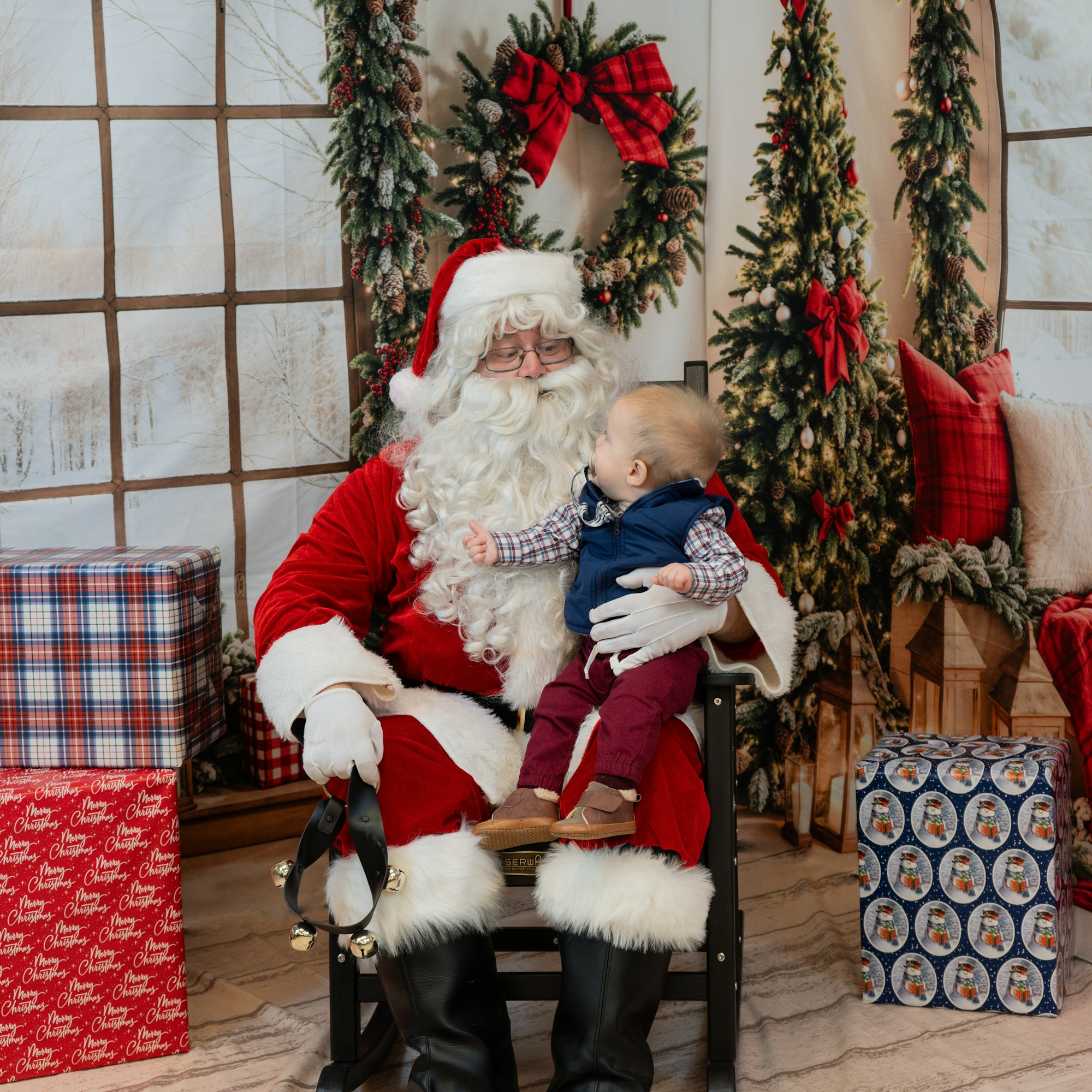 Santa holding a baby on his lap surrounded by Christmas decorations and gifts.