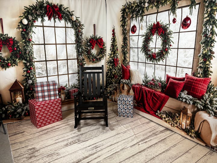 Cozy holiday scene; window, wreaths, gifts, black chair, red blanket, pillows, candle lanterns, and festive greenery.