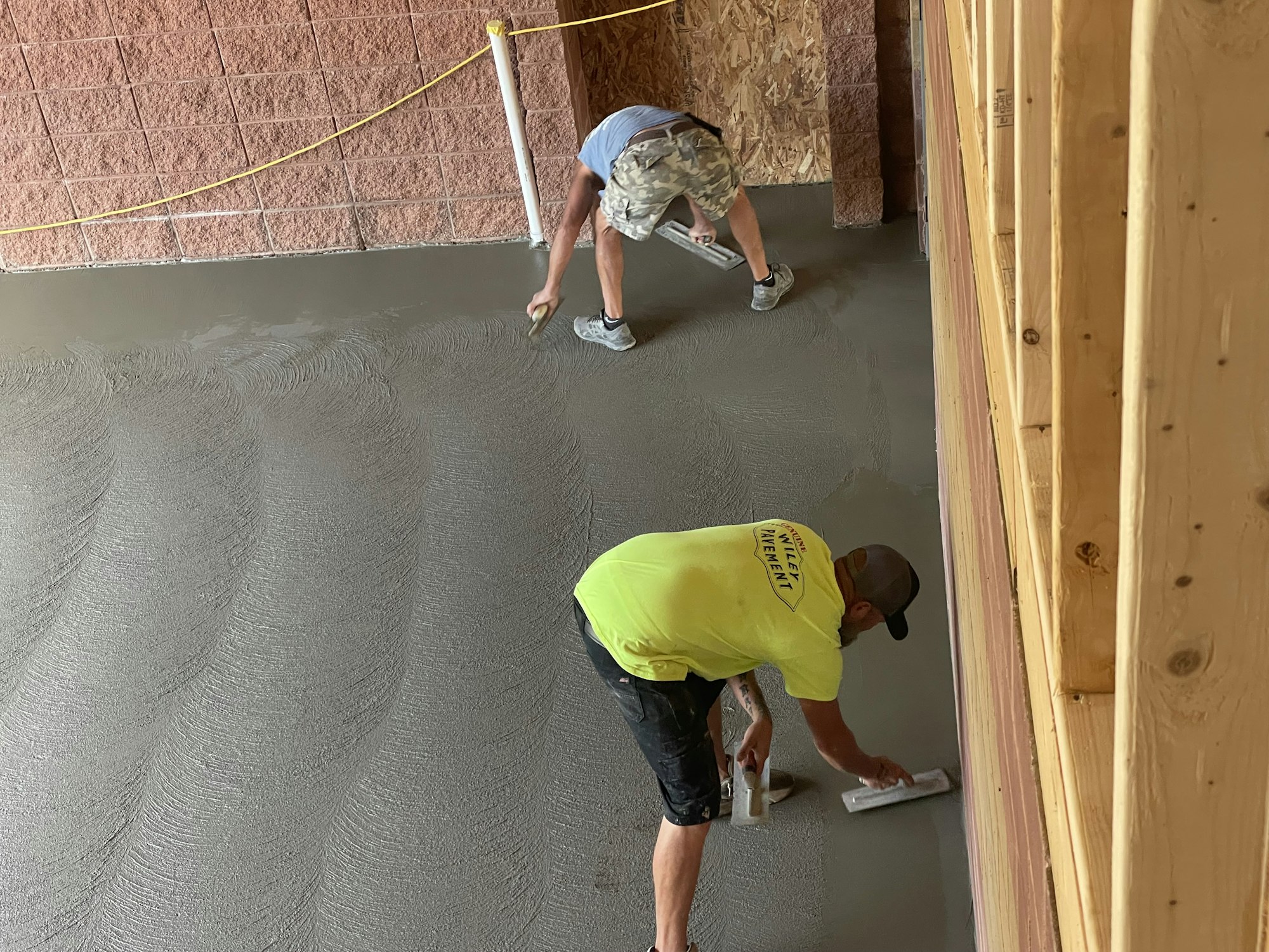 Two workers smoothing wet concrete on a floor indoors, with construction materials around.