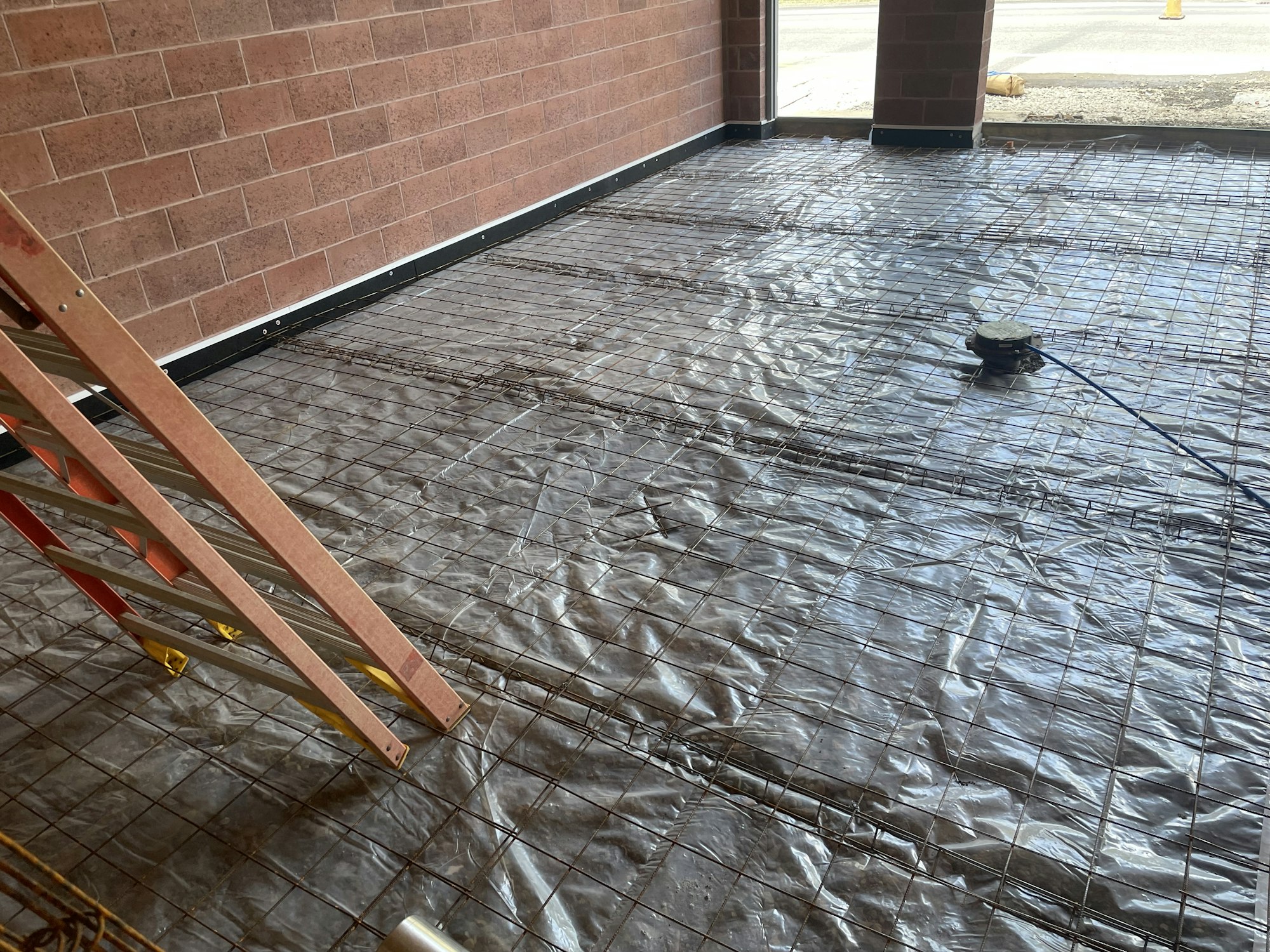 Construction site with rebar on a plastic sheet, likely prepping for a concrete pour, with a ladder to the left.
