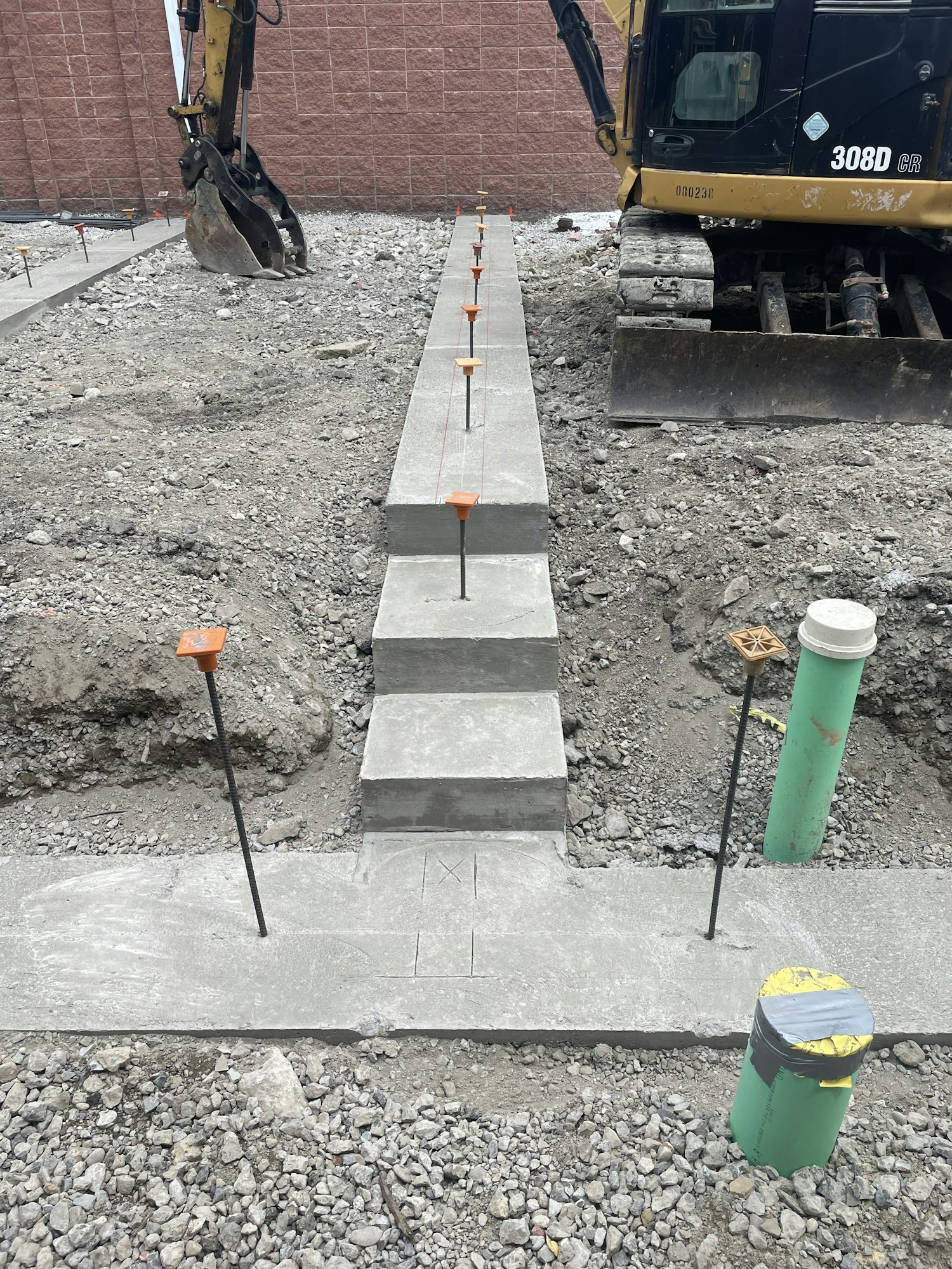 Construction site with a backhoe, newly laid concrete walkway slabs, and markers.