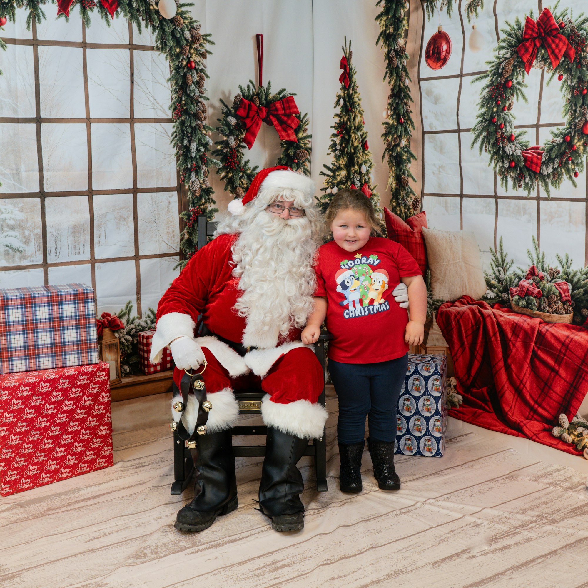 Santa with a child in a festive setting, surrounded by wreaths, trees, and wrapped gifts.