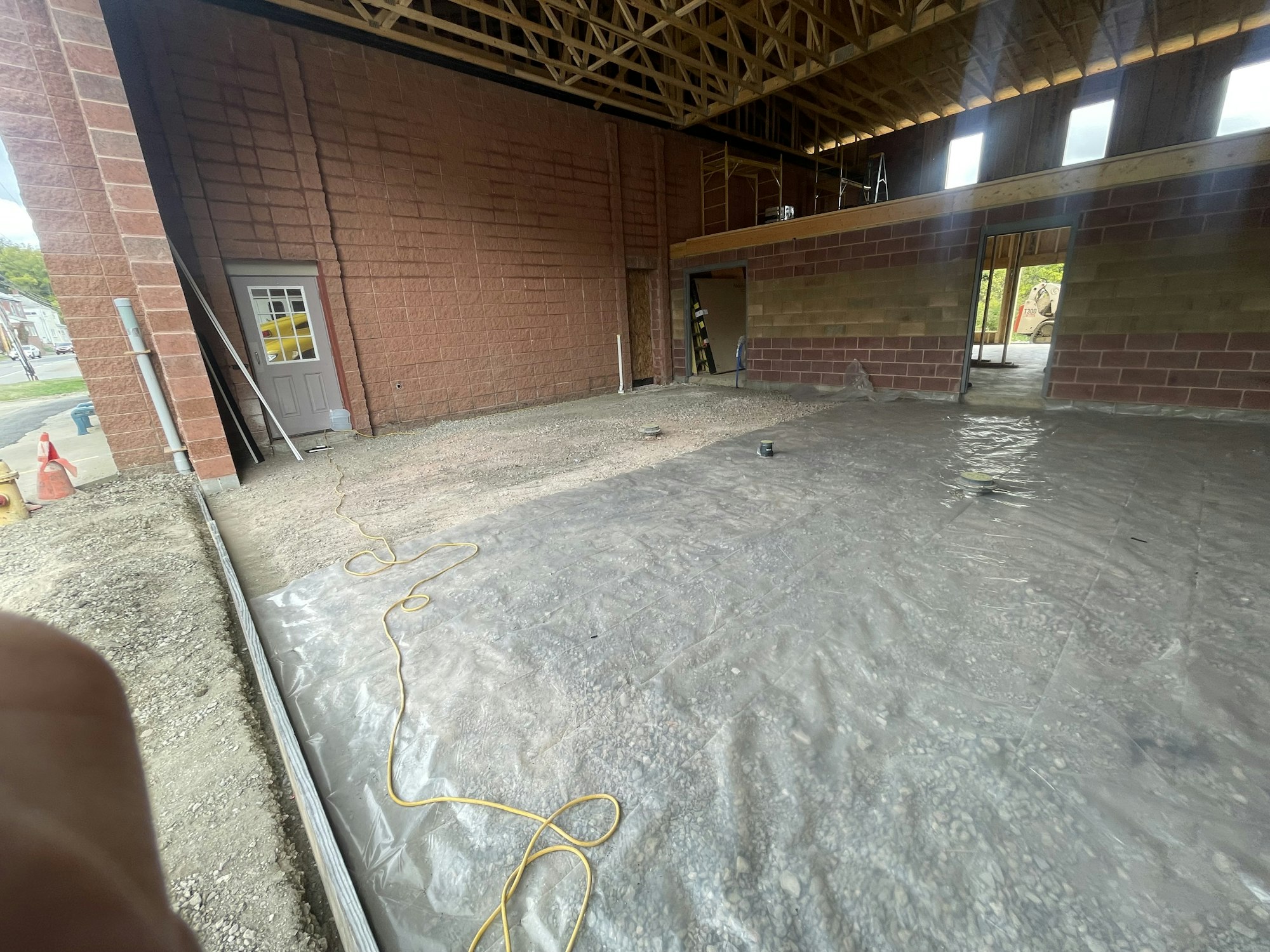 An unfinished interior space with exposed brick walls, concrete floor, wooden roof structure, doorways, and construction materials.