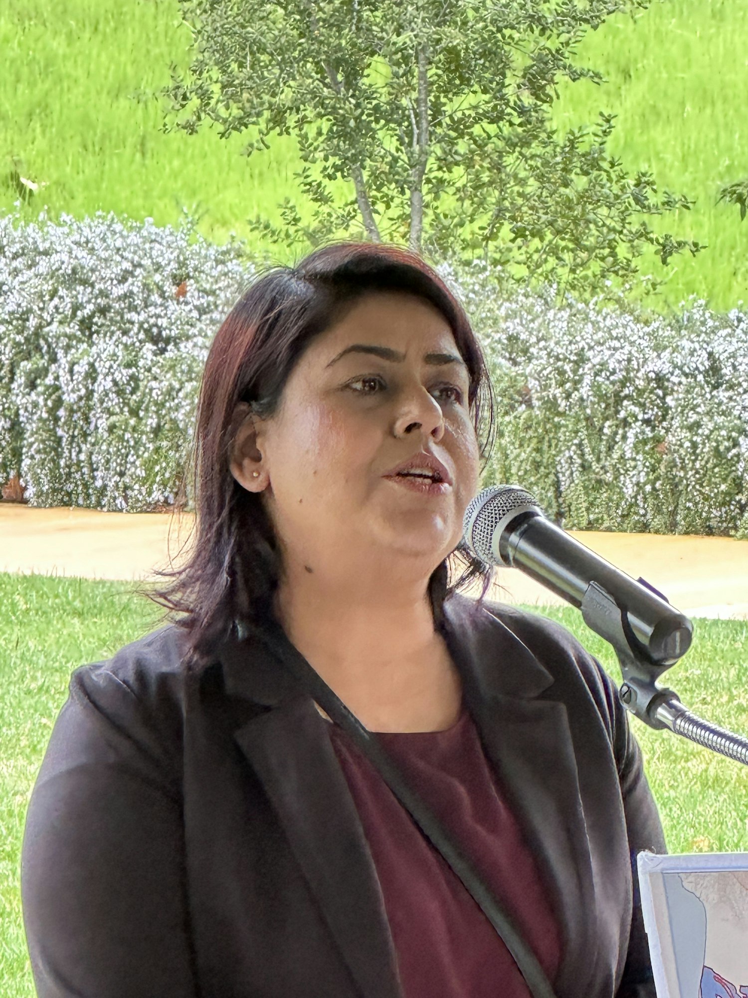 A woman is speaking into a microphone, with greenery in the background. (Note: The image is rotated.)