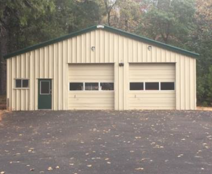 A large metal building with a green roof and two garage doors, surrounded by trees and a paved area.