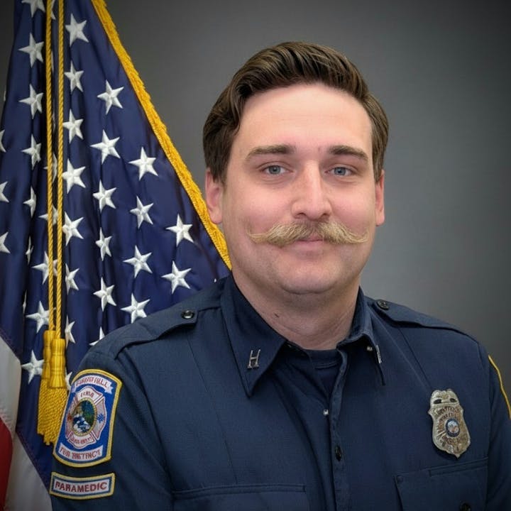 A smiling paramedic with a distinctive mustache poses in front of an American flag backdrop.