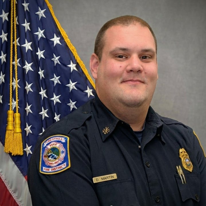The image features a man in a fire department uniform standing in front of an American flag backdrop.