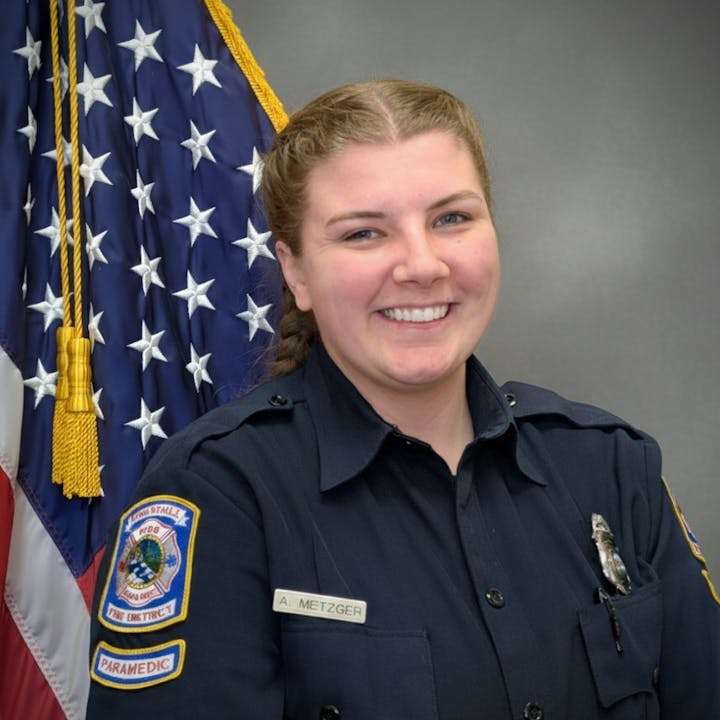 A smiling paramedic in uniform poses in front of an American flag backdrop.