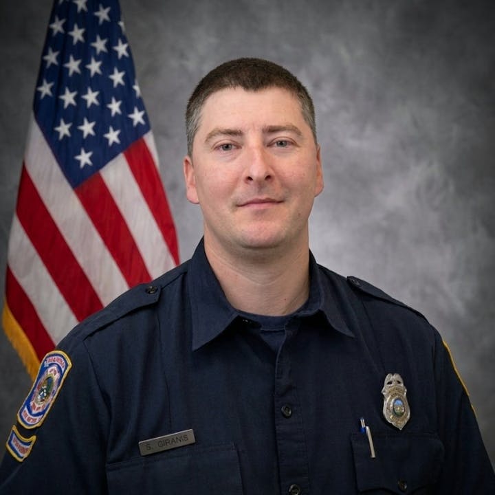 The image features a man in a dark uniform with a nameplate, standing in front of an American flag.