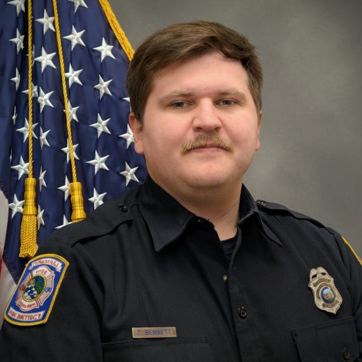 A uniformed firefighter poses in front of an American flag, displaying a badge and name tag labeled "Z Bennett."
