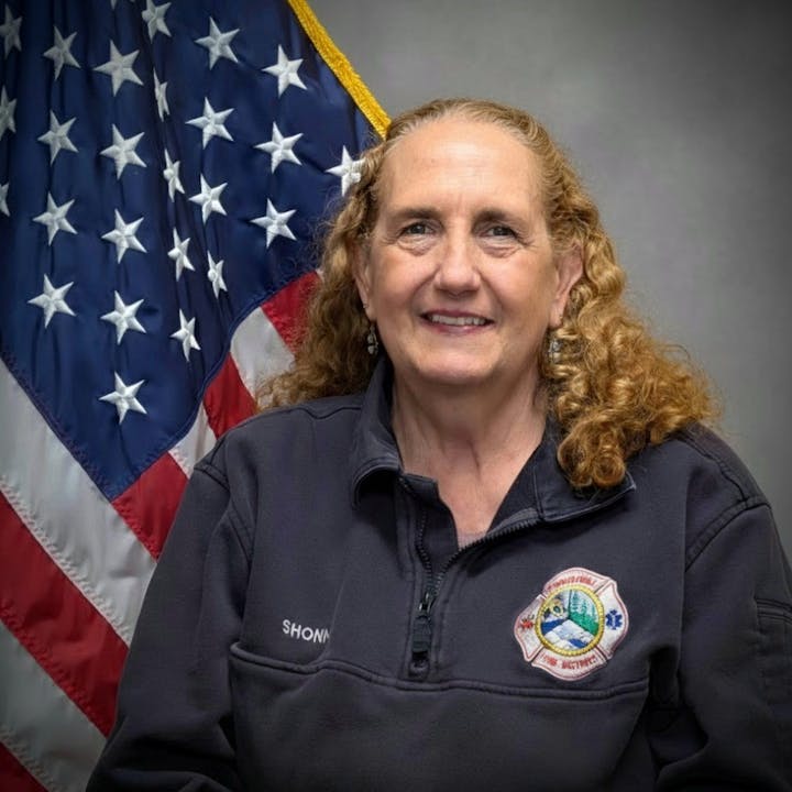 A smiling woman in front of an American flag, wearing a dark jacket with a fire department emblem.