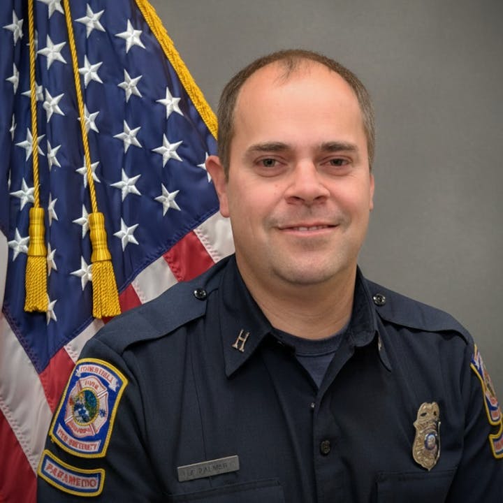 The image shows a man in a firefighter uniform standing in front of an American flag, smiling.