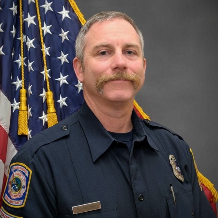 A uniformed individual stands in front of an American flag, sporting a distinctive mustache and a badge.