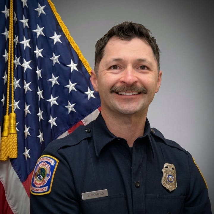 A smiling man in a uniform with a badge, standing in front of the American flag.