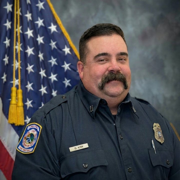 A man in a firefighter uniform smiles in front of the American flag. His nameplate reads "G. Kirk."