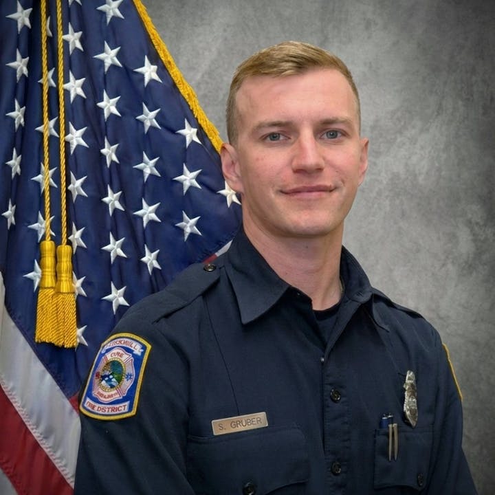The image features a smiling individual in uniform, standing in front of an American flag backdrop.