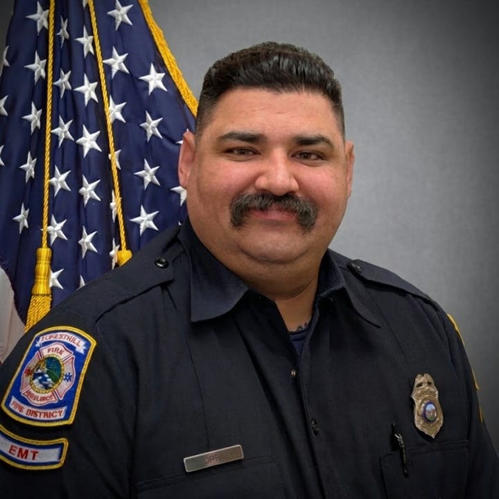 The image shows a uniformed emergency responder posing against a backdrop with an American flag. He has a mustache and a friendly expression.