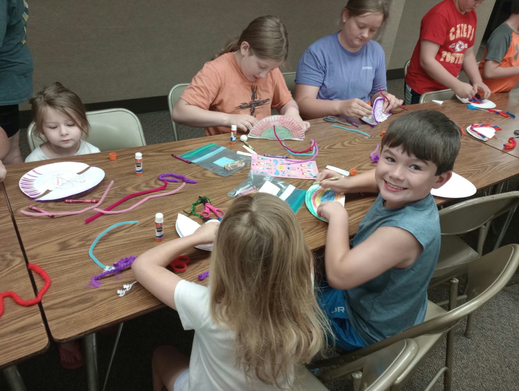 Children crafting at a table with paper plates, pipe cleaners, and glue sticks. A boy smiles at the camera.
