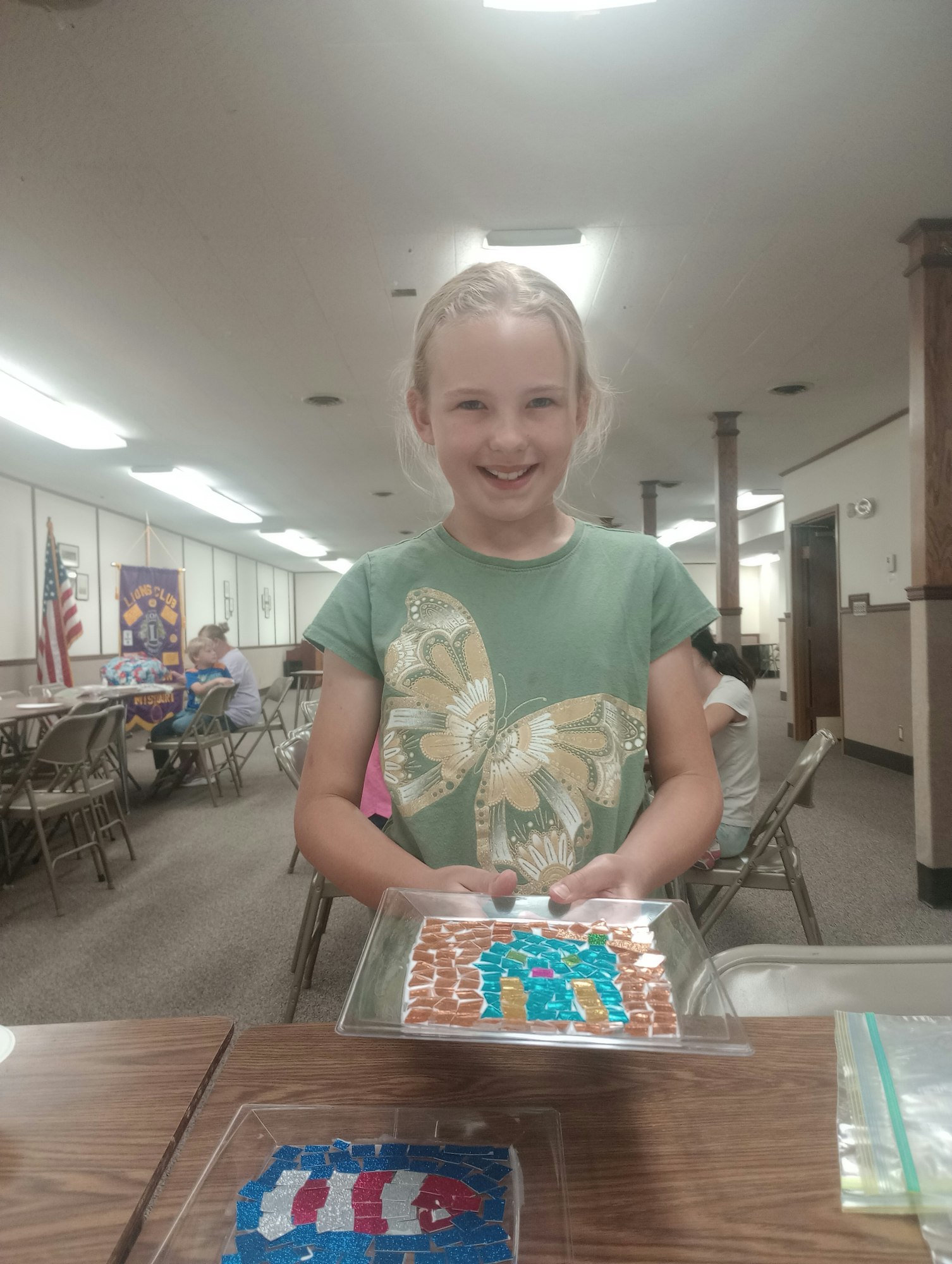 A child holds a mosaic art piece, smiling in a room with chairs and flags in the background.