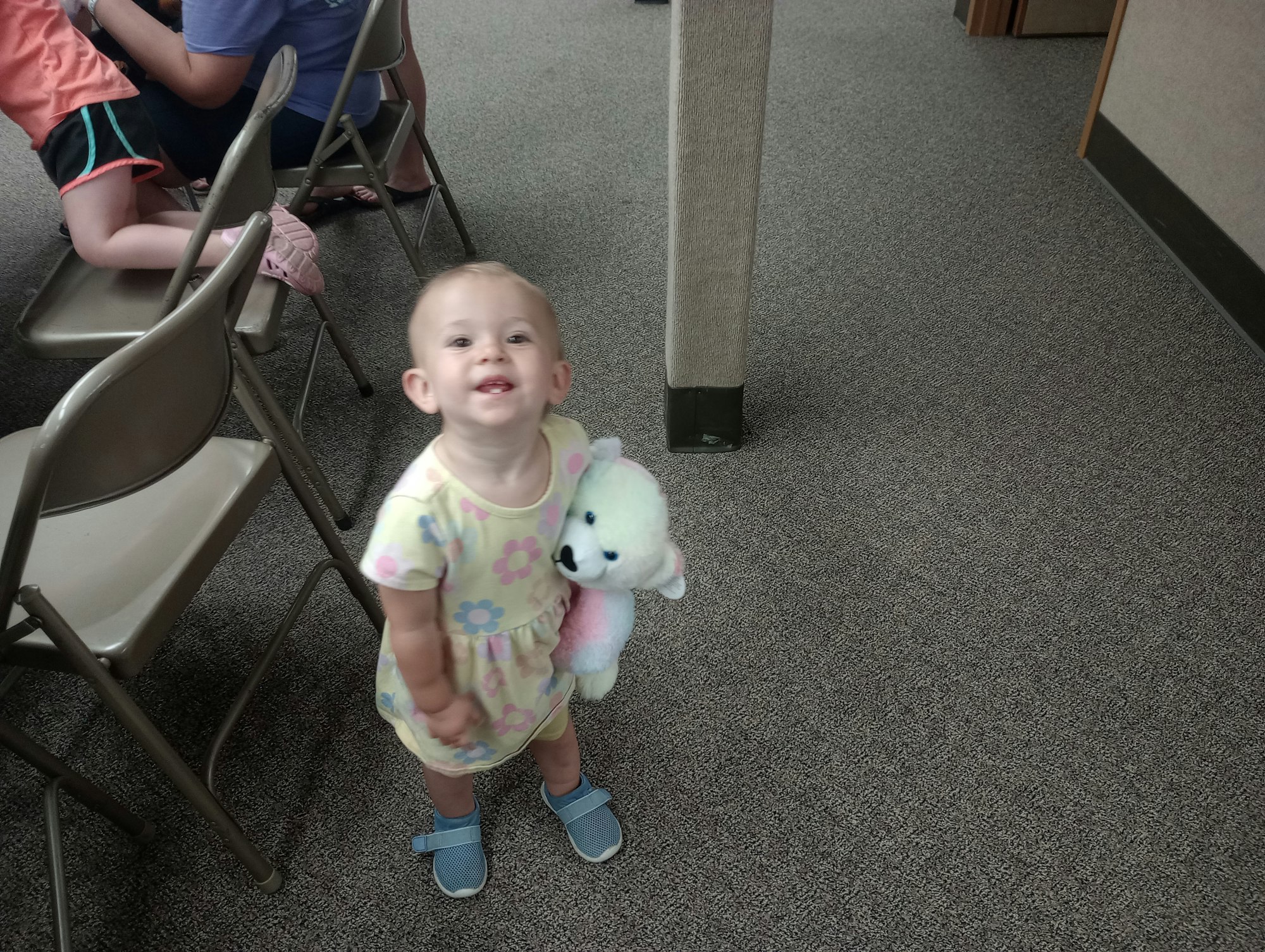 A smiling toddler holding a stuffed toy stands on a carpeted floor beside metal folding chairs.