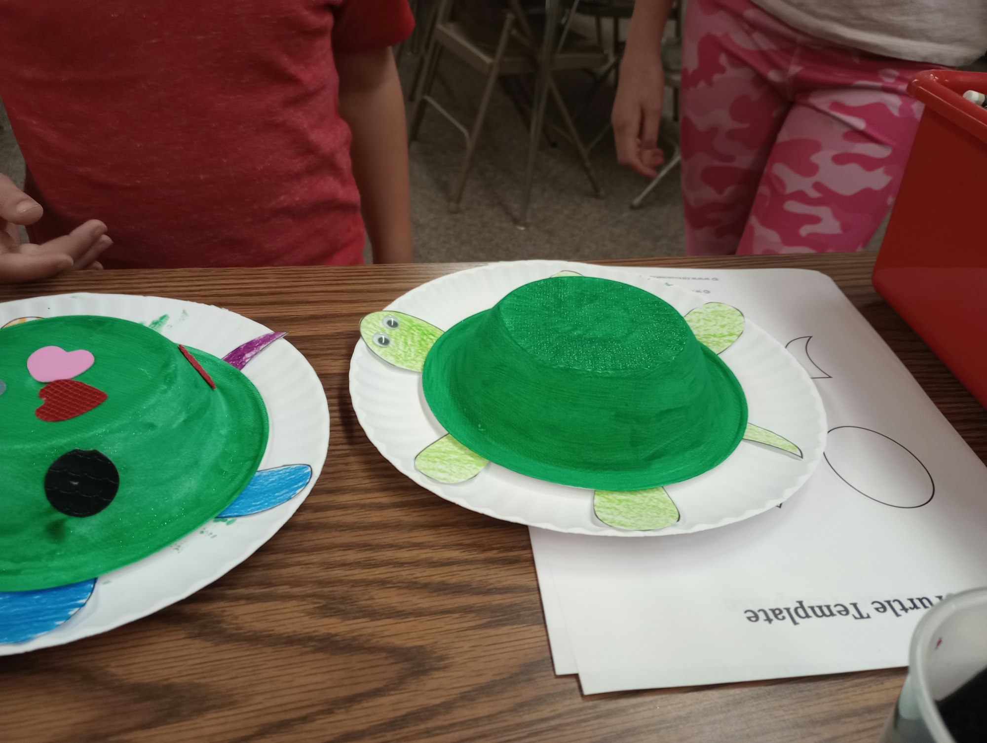 Two paper turtle crafts made from green painted bowls and plates are on a table, with kids nearby.
