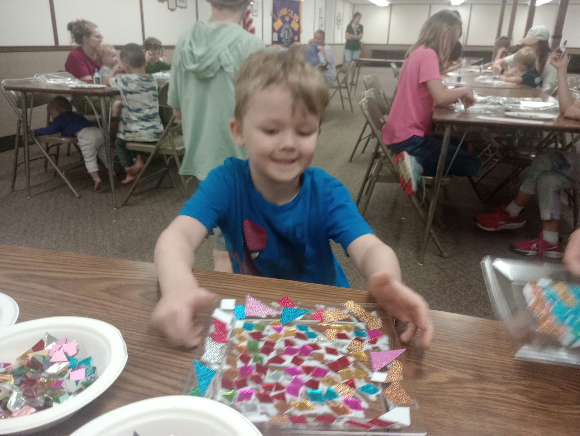 A child in blue shirt doing a colorful mosaic art project in a busy craft room with others working at tables.