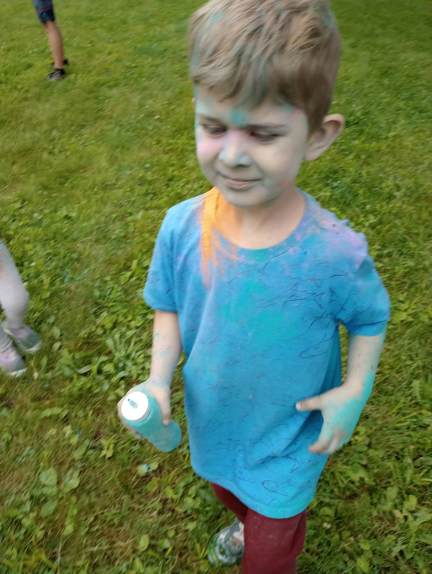 A child with colored powder on their shirt and face, holding a spray can, standing on grass.
