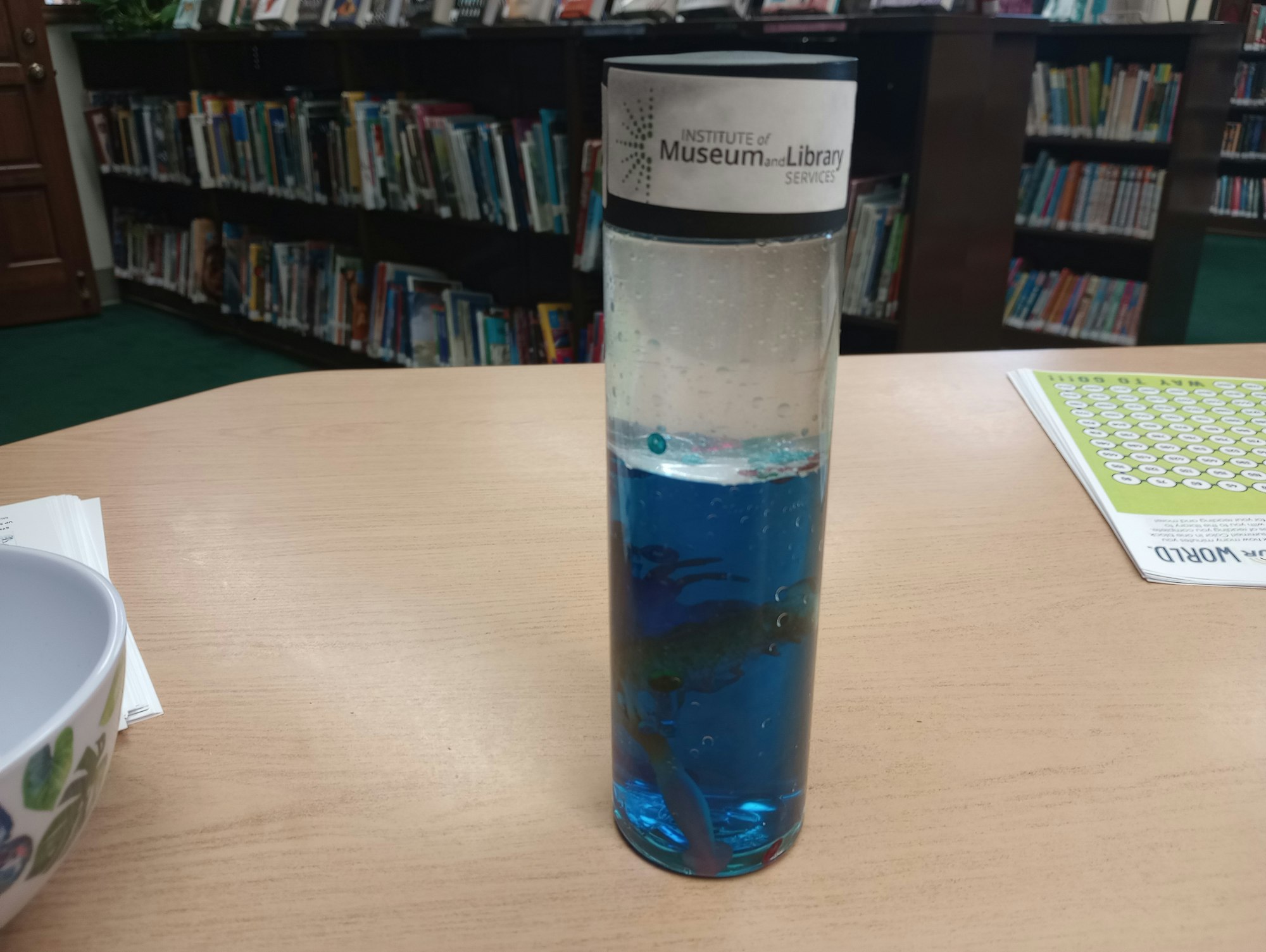 A cylindrical container with blue liquid and small objects inside, on a table in a library setting.