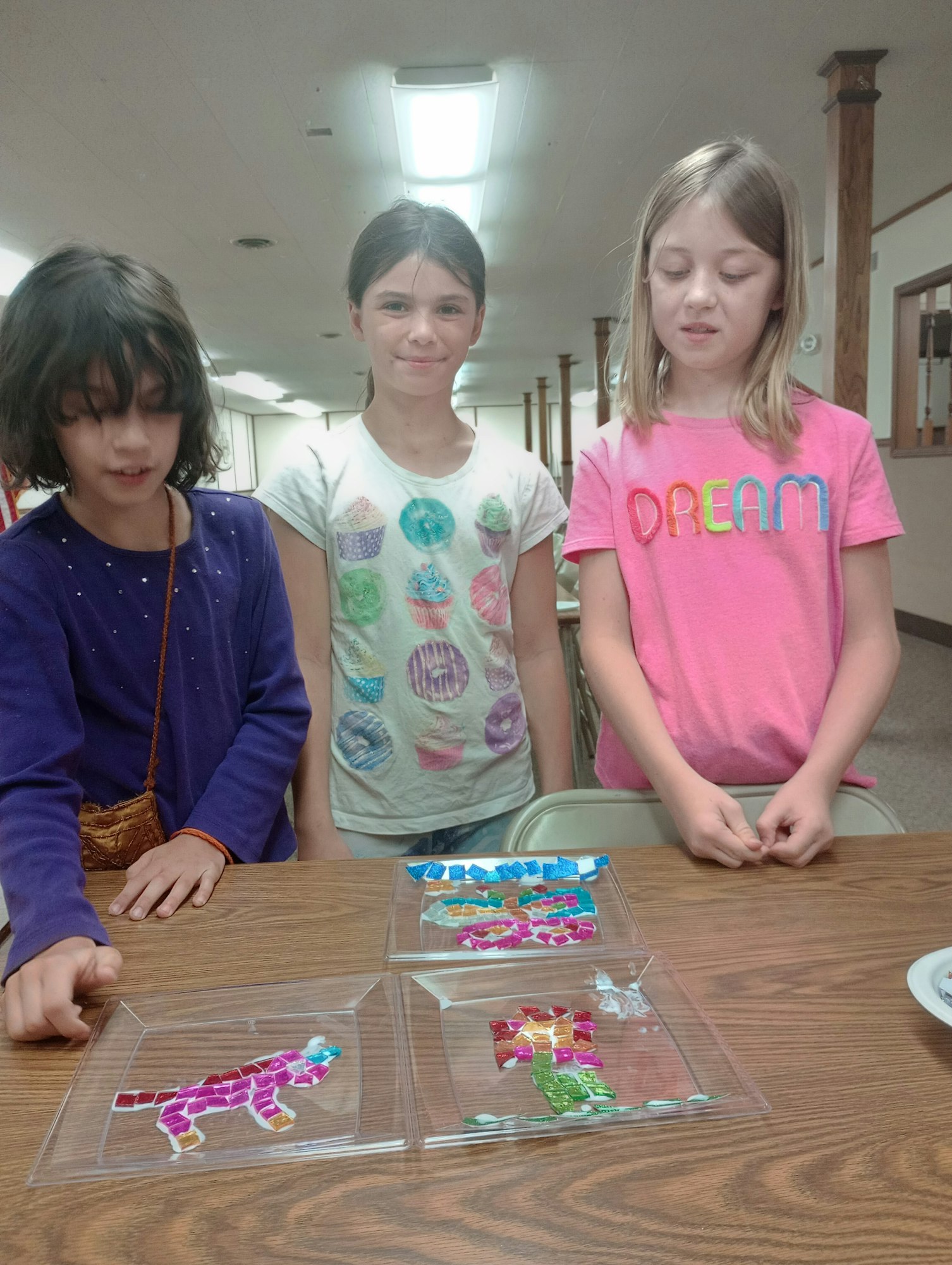 Three children standing at a table with colorful mosaic animal art pieces.
