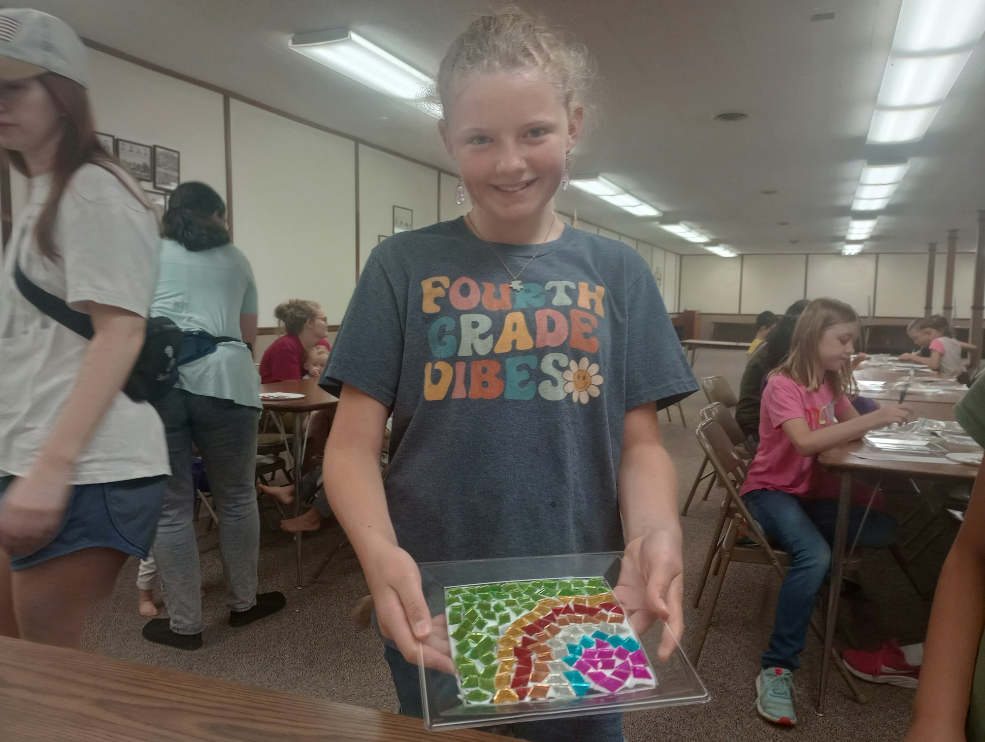 A smiling girl holds a colorful mosaic art piece, wearing a "Fourth Grade Vibes" shirt in a classroom with others working.