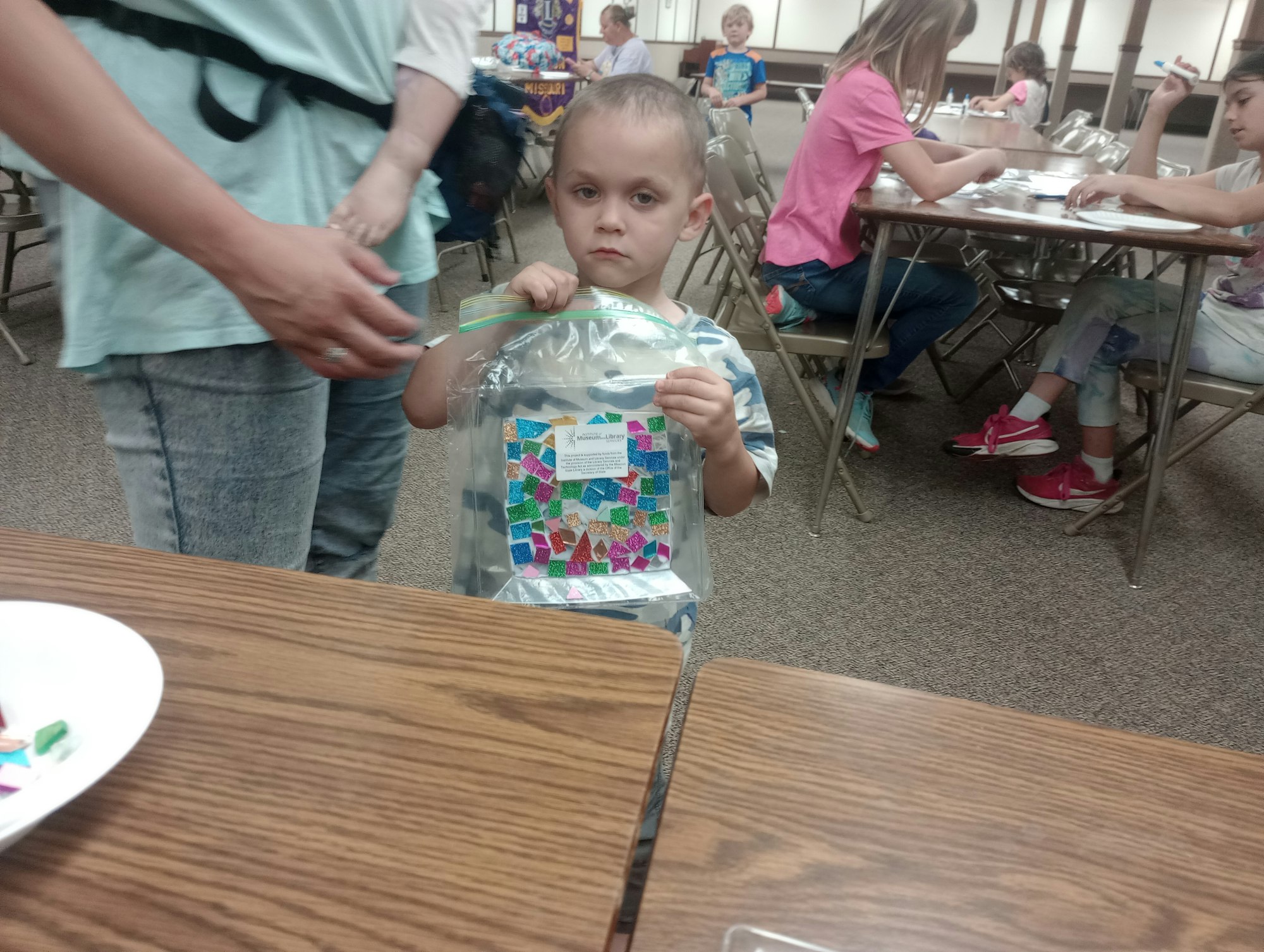 A child holding a decorated bag in a room with kids at tables doing crafts.