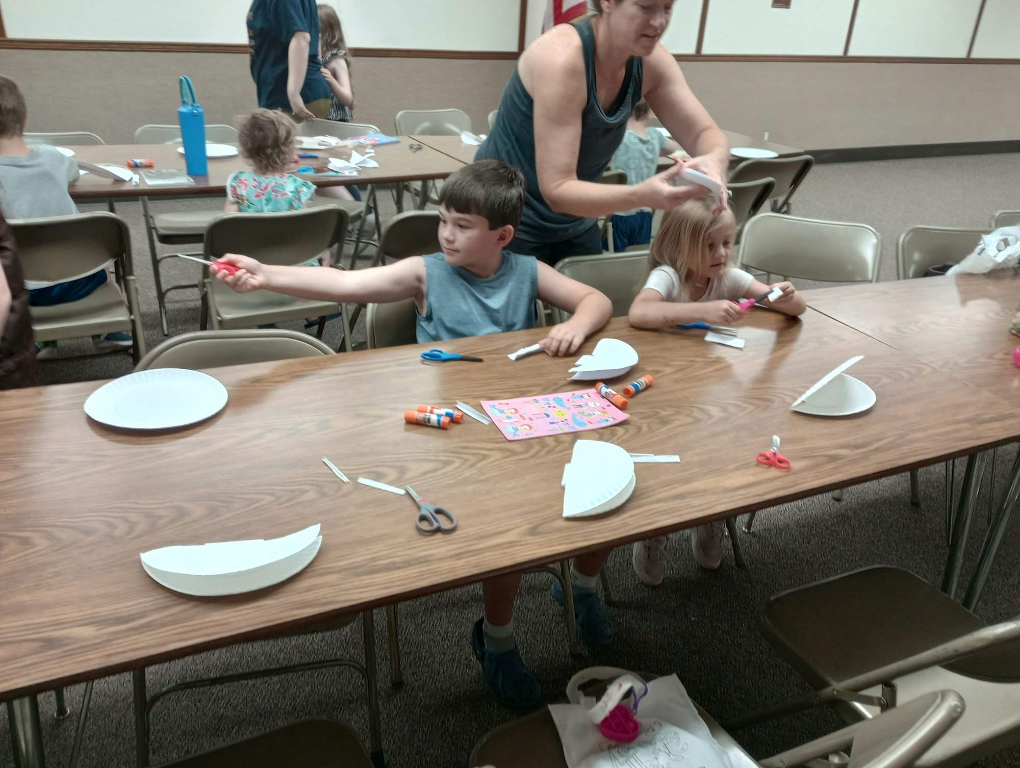 Children and an adult engaged in a craft activity with scissors, paper plates, and glue sticks on a table.