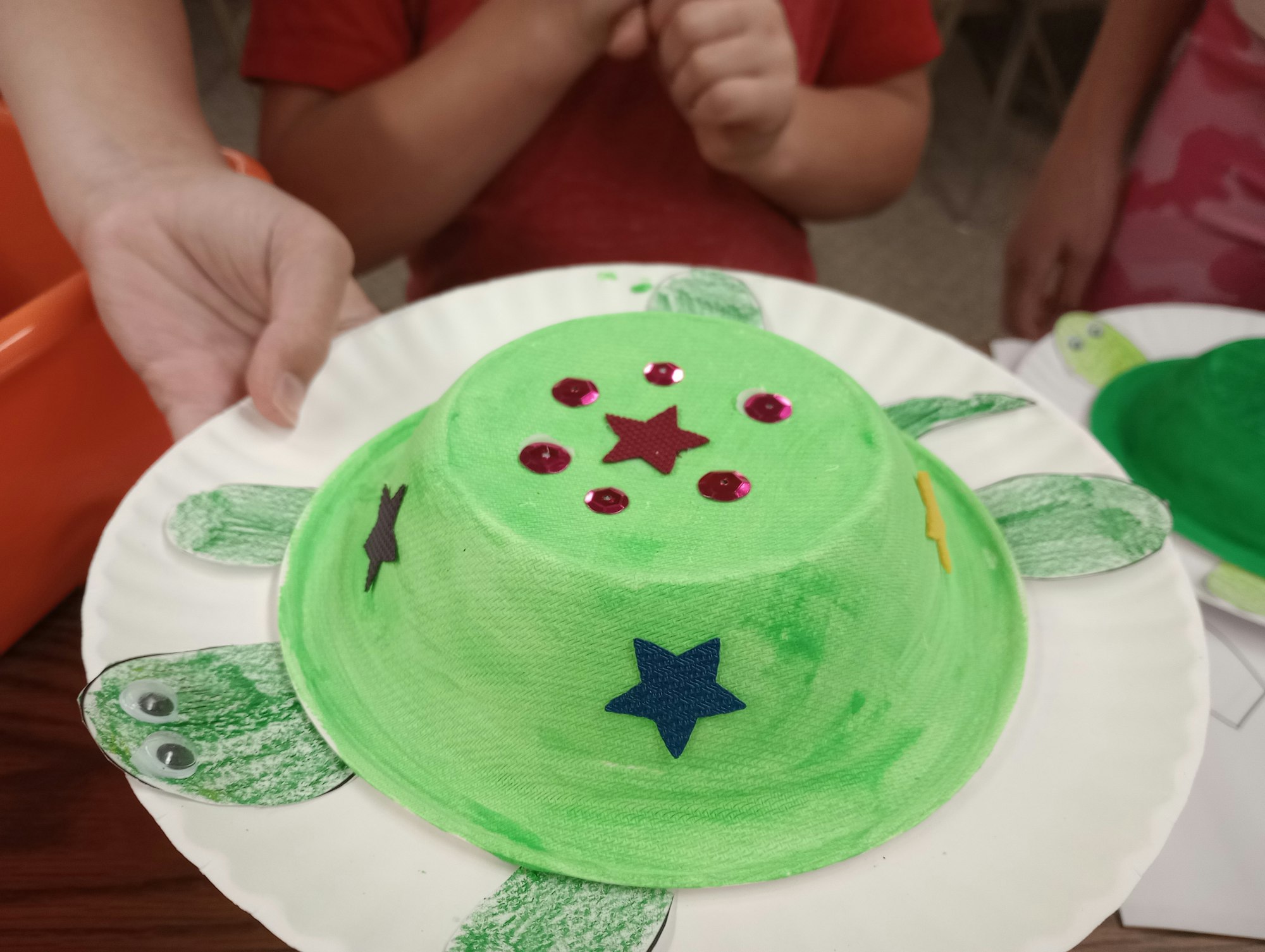 A child holds a paper turtle craft, decorated with stars and sequins, on a paper plate.