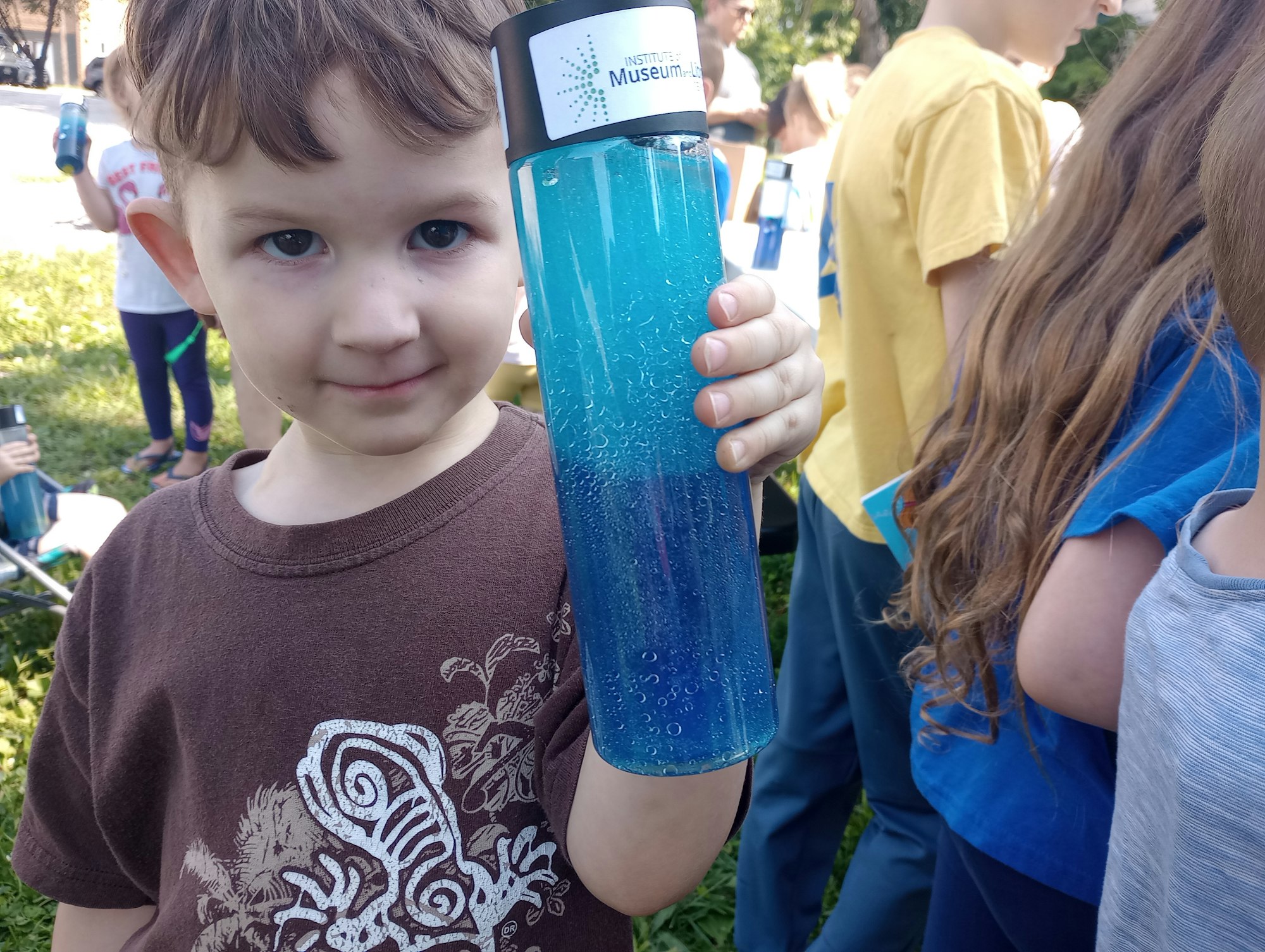 A child holding a blue bottle with "Museum" on it, surrounded by other children outdoors.