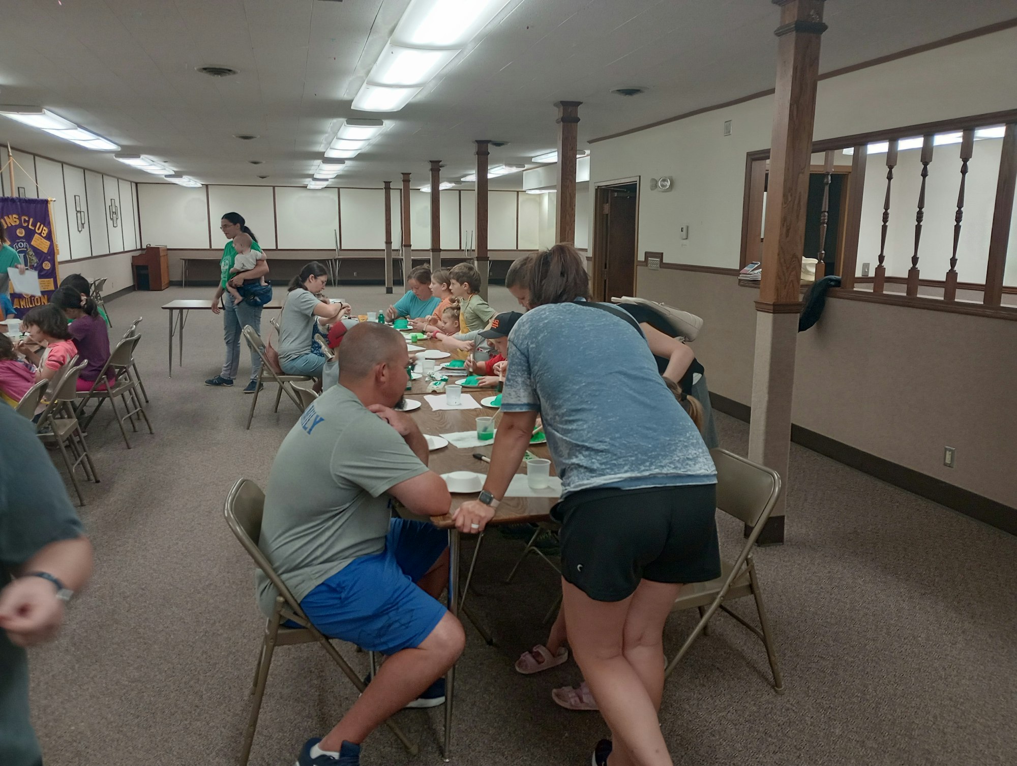 People are gathered in a room with tables, eating or socializing, under bright ceiling lights.
