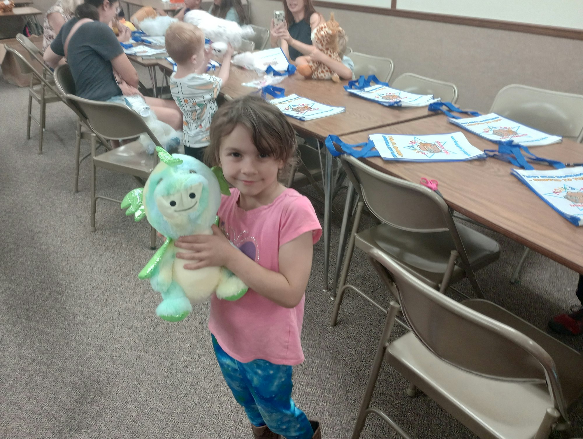 Child holding a green stuffed toy, with people crafting at a table in the background.