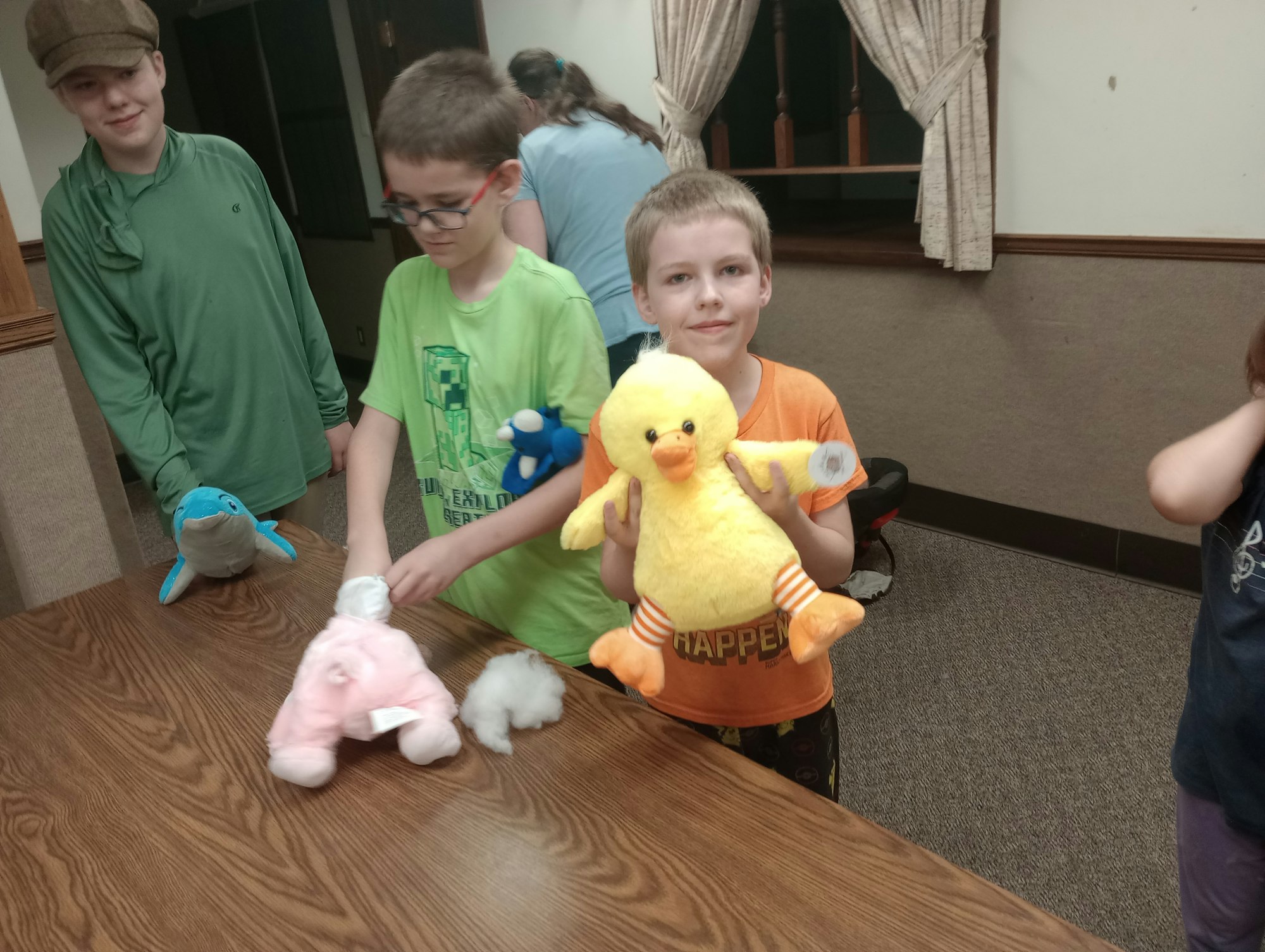 Kids with stuffed animals around a table, one holding a yellow duck plush toy.