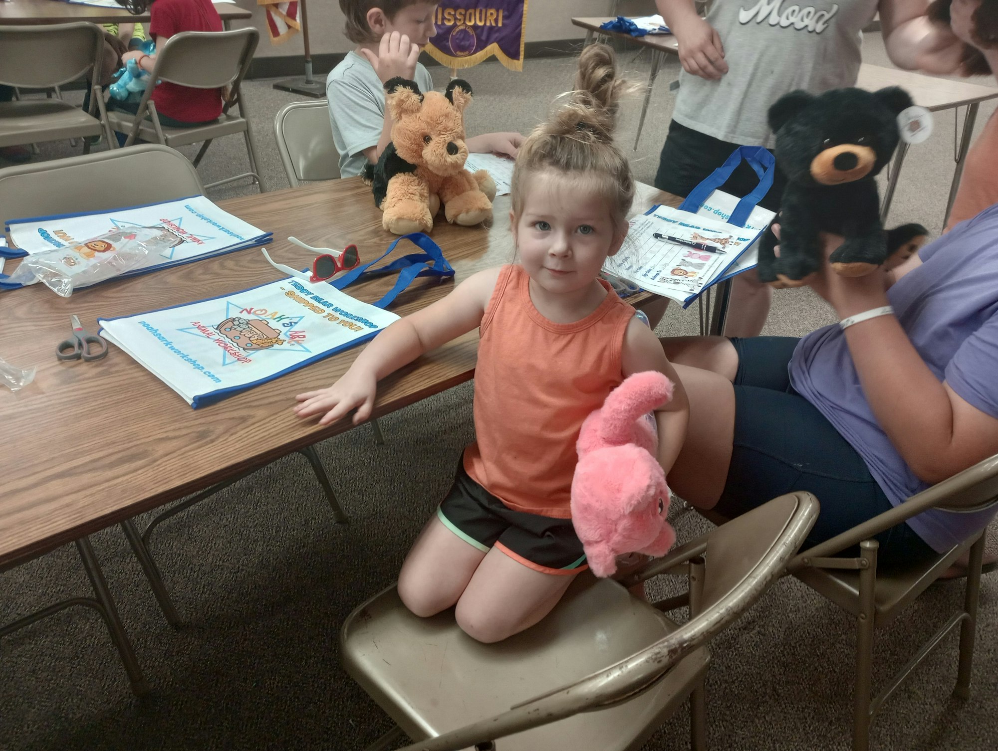 A child holding a pink stuffed animal at a table, with bags and teddy bears around. Other people are seated nearby.
