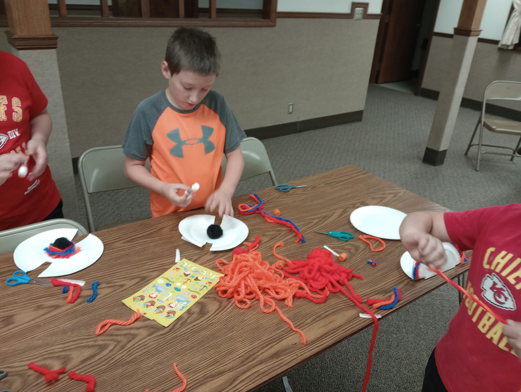 Children crafting with paper plates, yarn, scissors, and glue sticks on a table.