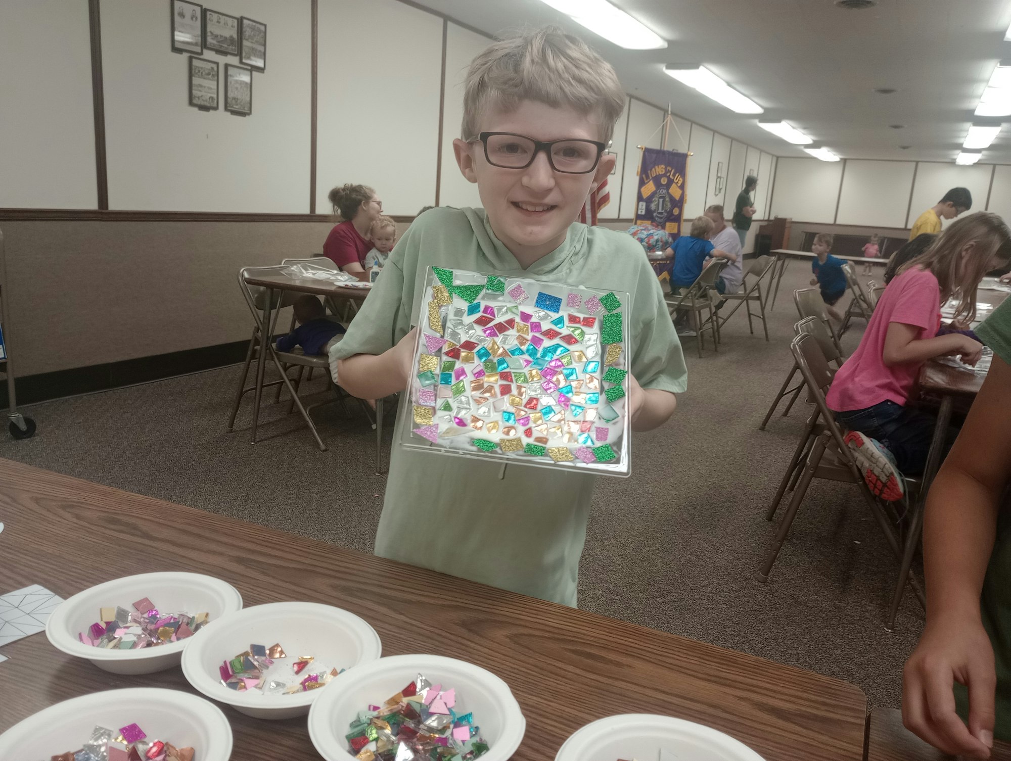 A child with glasses holds a colorful artwork made of shiny shapes in a room with others doing craft activities at a table.