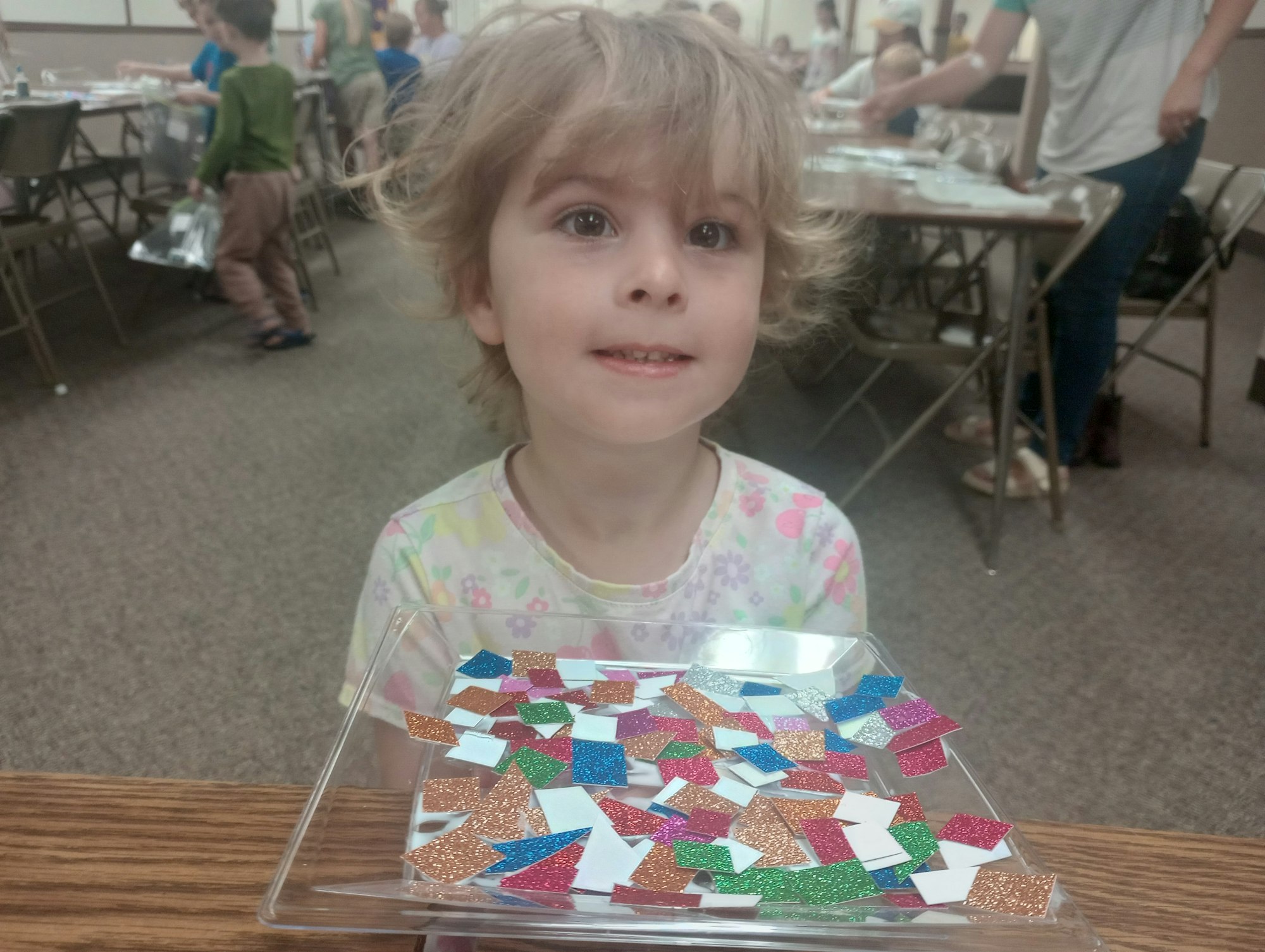A child holds a tray with colorful glitter paper pieces in a room with tables and people in the background.