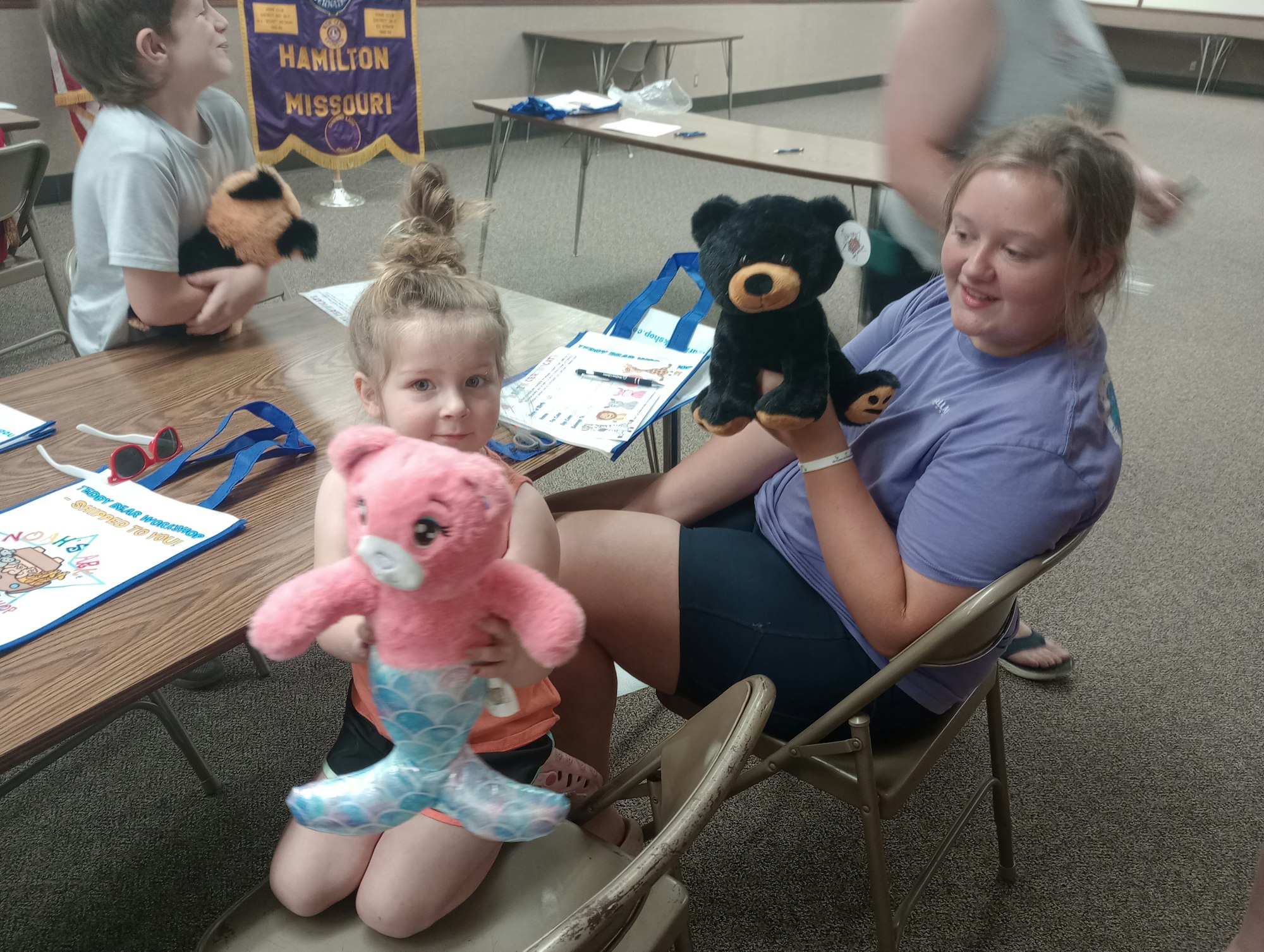 Kids holding plush toys, sitting at a table with activity sheets, and a "Hamilton Missouri" banner in the background.