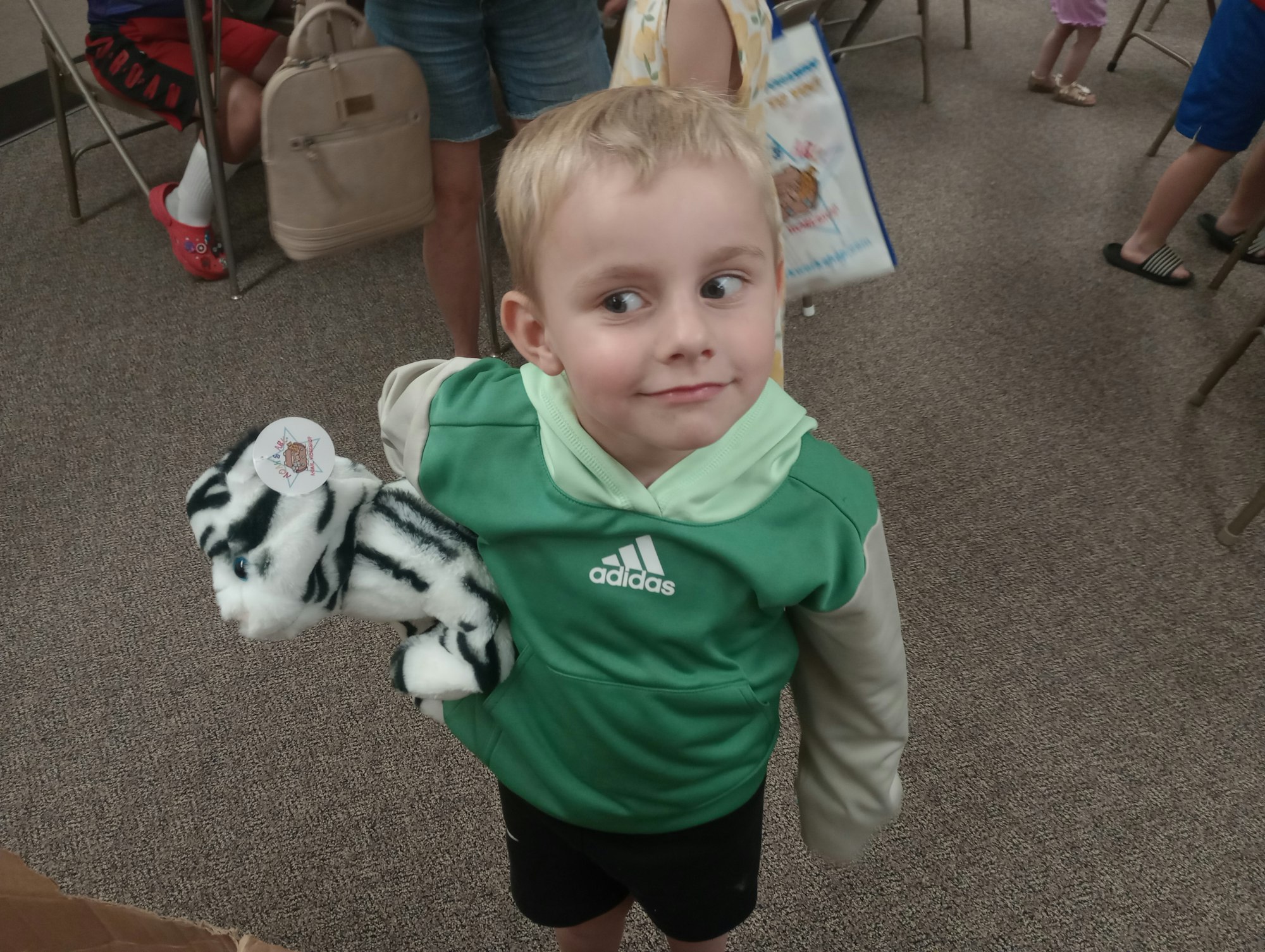 A child in a green jacket holding a stuffed zebra toy, surrounded by people and chairs.
