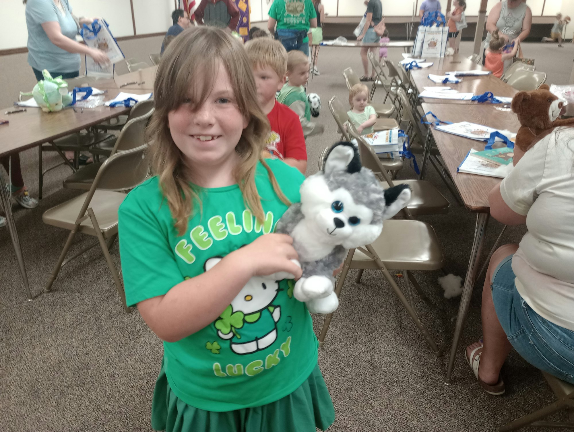 A child in a green shirt holding a stuffed animal in a room with other children and tables.