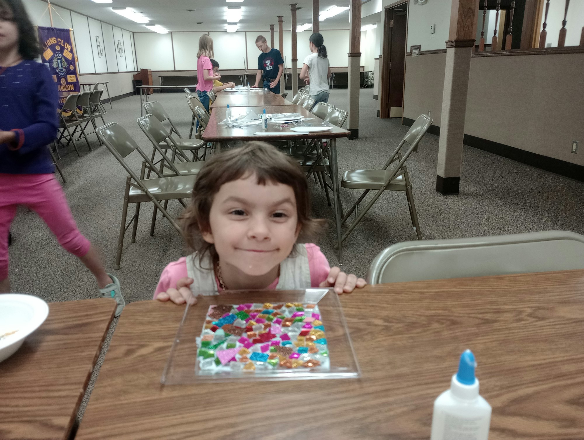 A child smiling at a table with a colorful mosaic piece, glue bottle nearby; several children in the background in a hall.