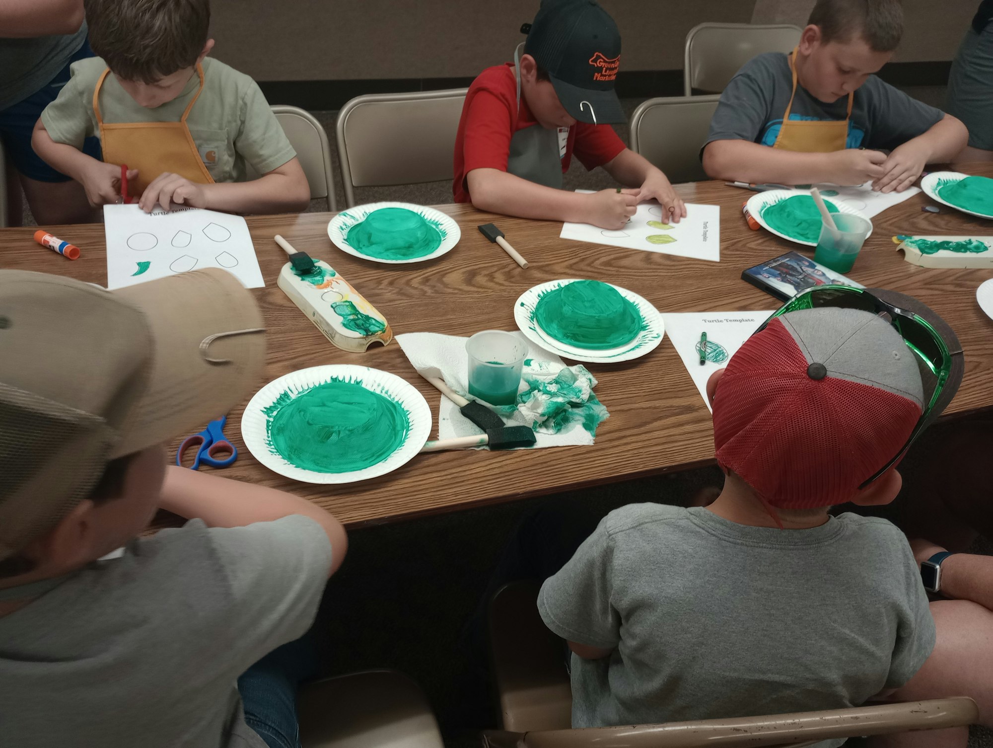 Kids are doing an art activity with green paint, paper plates, and templates on a table.