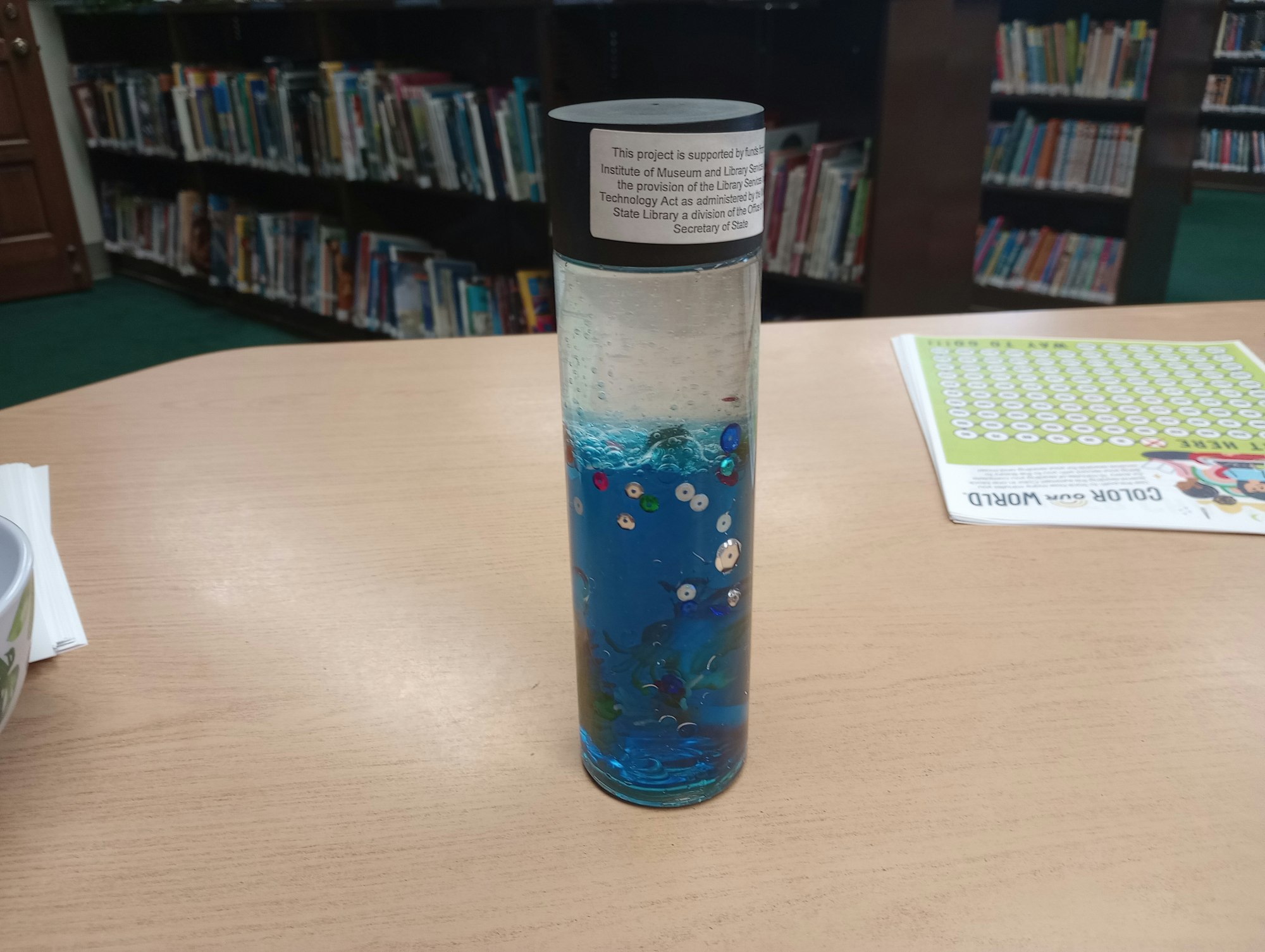 A blue sensory bottle with sequins on a table in a library.